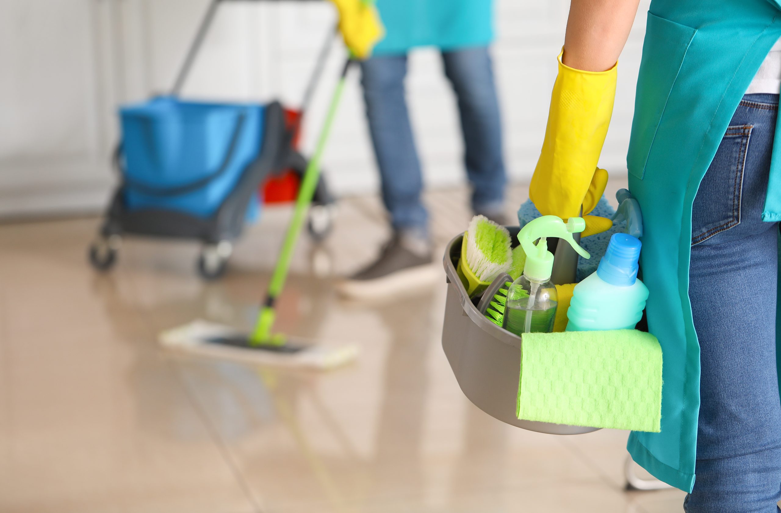 Two people cleaning a floor; one holds a mop, the other carries a caddy with cleaning supplies and wears yellow gloves.