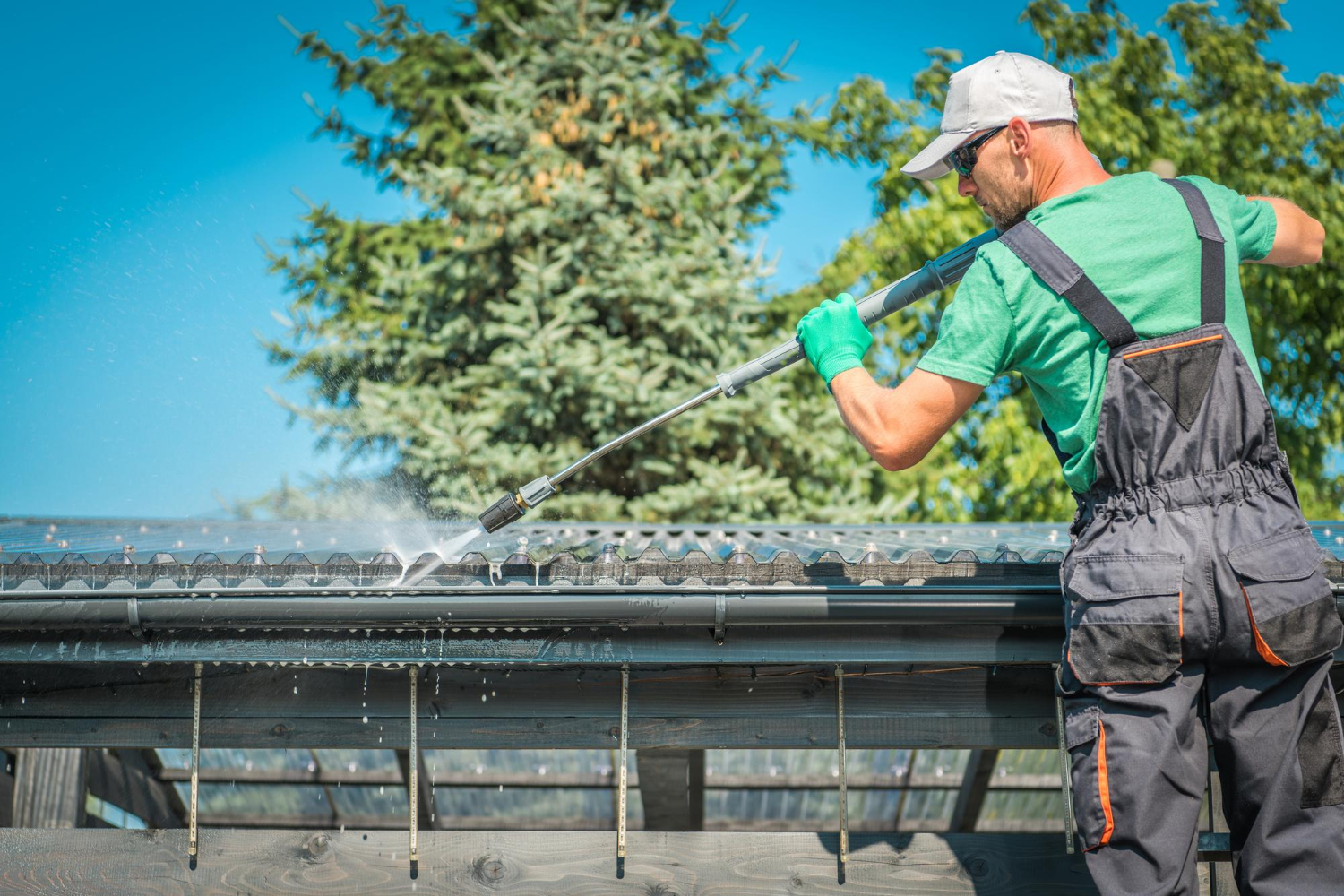 Man in work clothes pressure washing a roof gutter outdoors on a sunny day.