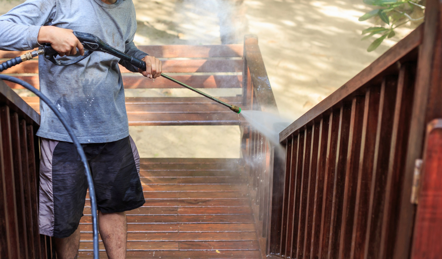 Person using a pressure washer to clean wooden outdoor stairs on a sunny day.