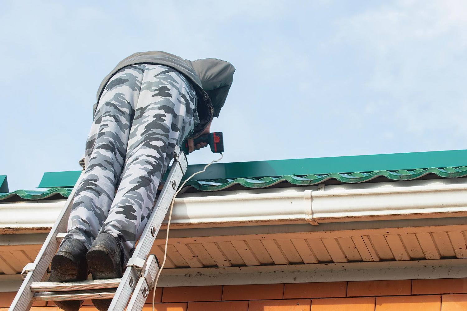 Person in camouflage pants using a power drill on a green roof while standing on a ladder.